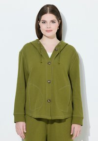 Olivově zelená bunda s kapucí s texturovaným povrchem, která má pět knoflíků, dvě velké kapsy a nastavitelná šňůrky na kapuci.