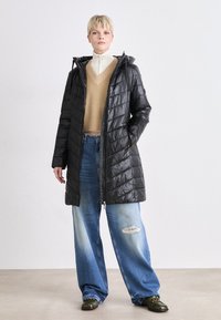 Kvinne med kort blond hår iført en lang svart pufferjakke, beige V-hals genser, vide ripped jeans, og grønne sko, stående.