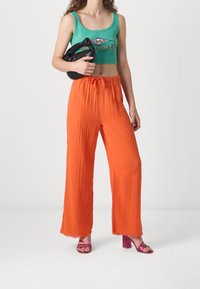 Crop top vert avec logo, pantalons larges orange en tissu texturé, et sandales à talons roses. Le mannequin tient un sac noir.