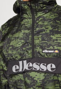 Grøn camouflage vindjakke med et sort panel, der prydes af "ellesse" logoet i hvid. Struktureret stof med høj krave og lynlås.