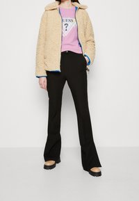 Beige, huňatá bunda na zip s modrým lemem přes růžové tričko s logem. Černé kalhoty do zvonu a béžové platformy dotvářejí outfit.