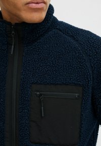 Marine fleece jack met een gestructureerde stof, hoge kraag en zwarte nylon ritszak op de borst. Volledige ritssluiting en ribbelige zijkanten.