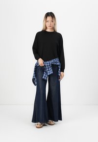 Zwarte lange mouwen shirt, hooggewaisted flared blauwe spijkerbroek en een blauw geruit shirt om de taille geknoopt. Beige schoenen met accenten.