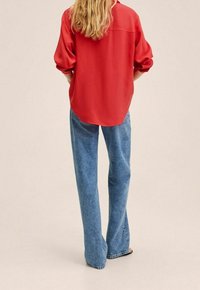 Persona con lunghi capelli biondi, indossa una maglietta rossa a maniche lunghe e jeans blu larghi, in piedi con le mani in tasca, di spalle.