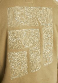 Tissu beige avec une broderie abstraite blanche formant un motif rectangulaire irrégulier avec un travail de boucle dense.