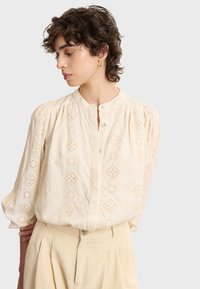 Crème-kleurige blouse met geborduurde diamanten en bloemmotieven, voorzien van een ronde halslijn, knopenlijst en puffmouwen.