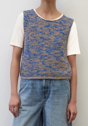 Personne portant un gilet tricoté bleu et beige par-dessus une chemise blanche à manches courtes et un jean en denim bleu clair, debout contre un mur uni.