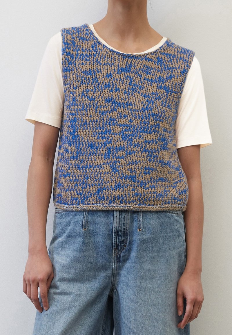 Personne portant un gilet tricoté bleu et beige par-dessus une chemise blanche à manches courtes et un jean en denim bleu clair, debout contre un mur uni.