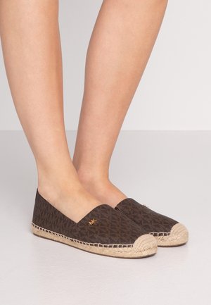 Stopy w brązowych klapkach espadrylach z monogramem, posiadających beżowe skręcane noski i podeszwy, stojące na szarej podłodze.