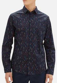 Chemise bleu marine à boutons avec un motif géométrique orange, blanc et rouge. Elle présente un col pointu et une poche sur la poitrine gauche.