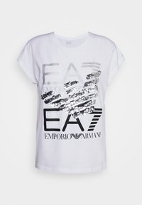 Vit t-shirt med kort ärm som har silver- och svart text med paljetter i en grafisk design. Texten lyder "EA7 EMPORIO ARMANI."