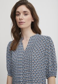 Modrá a bílá halenka s vzorem, V-výstřihem, sklady a balónovými rukávy; geometrický design obsahuje střídající se čáry a tvary na tmavém pozadí.