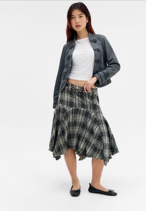 Jeune femme portant un top court blanc, une veste boutonnée gris foncé, une jupe asymétrique à carreaux noir et blanc, et des ballerines noires, debout sur un fond blanc.