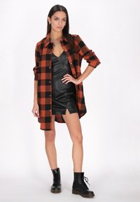 Camicia in flanella a quadri rossi e neri, stile button-down, sopra un vestito attillato in pelle nera con uno spacco anteriore, abbinato a stivali neri con lacci.