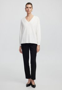 Bílý svetr s výstřihem do V a dlouhými rukávy, doplněný černými střiženými kalhotami. Texturovaná tkanina s uvolněným střihem; celkový vzhled doplňují špičaté černé boty.