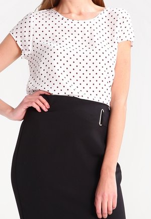 Vrouw draagt een wit topje met korte mouwen en zwarte polkadots, ingestopt in een hooggetailleerde zwarte rok met een zijkant gesp detail.