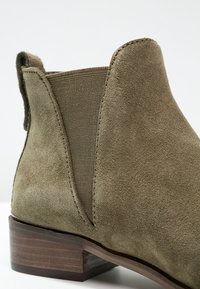 Bottine en daim vert olive avec des panneaux latéraux élastiques et un talon en bois empilé, présentant une texture lisse et des lignes épurées.