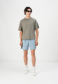 Grå oversized t-skjorte og lyseblå shorts. Glatt bomullsstoff. Svart solbriller. Hvite sneakers med teksturerte såler. Avslappet antrekk.