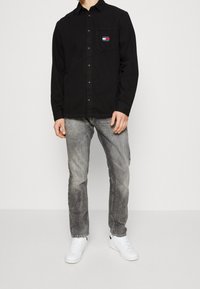 Camisa negra de manga larga con cierre de botones y un bolsillo en el pecho que lleva un logotipo en rojo, blanco y azul. Combinada con jeans grises desgastados y zapatillas blancas.