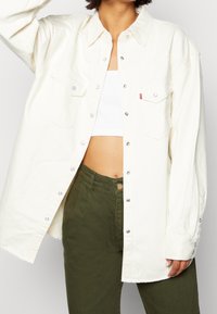 Mujer llevando una camisa abotonada de gran tamaño en color blanco roto sobre un top corto blanco y pantalones de tiro alto en verde olivo, con la mano descansando a un lado.