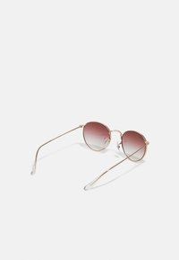 Kulaté sluneční brýle se zlatým rámem, gradientní hnědé čočky a průhlednými špičkami na nožičkách. Mají elegantní, minimalistický design.
