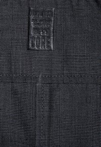 Černá textilie s kostkovaným vzorem, zdobená texturovanou černou koženou značkou s raženým nápisem „CORE“ a dalšími detaily značky.