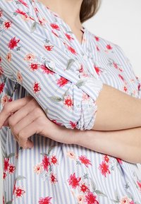 Blå och vit randig blus med röd och rosa blommig broderi. Har knäppte ärmslut och en avslappnad passform. Mjuk, lättviktig tyg.