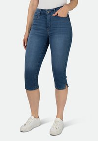 Denim Capri-Jeans in mittlerem Blau mit einem taillierten Design, Seitenschlitzen am Saum, fünf Taschen und einem Knopfverschluss. Getragen mit weißen Sneakers.