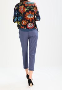 Kvinna i blå cropped byxor, svarta högklackade skor och en svart tröja med stort färgglatt blommönster, stående med ryggen mot betraktaren.