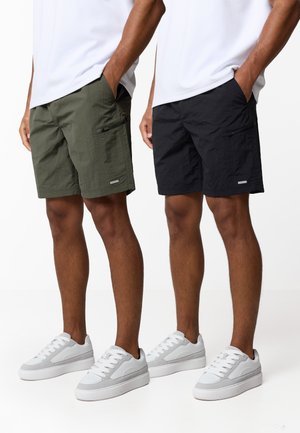 Δύο άνδρες φορούν λευκά αθλητικά παπούτσια και casual σορτς· ο ένας σε ελιά πράσινο με τσέπη με φερμουάρ, ο άλλος σε μαύρο, και οι δύο με τα χέρια στις τσέπες.