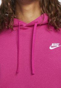 Růžová mikina s kapucí a bílým logem Nike na hrudi. Materiál vypadá měkký a hladký na dotek.