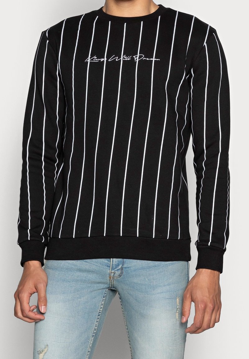 Chemise noire à manches longues avec des rayures verticales blanches et un texte blanc en écriture cursive sur la poitrine, portée avec un jean bleu clair délavé.