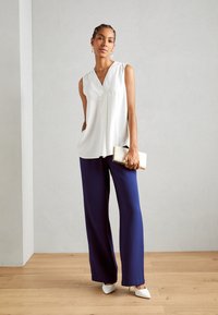 Blusa bianca senza maniche con scollo a V, abbinata a pantaloni larghi blu navy. Il modello tiene una pochette bianca e indossa décolleté bianchi a punta.