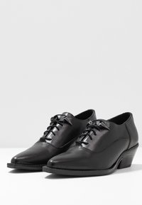 Chaussures en cuir noir avec bouts pointus, design à lacets, petit talon empilé et texture lisse. Détails subtils de couture le long des côtés.