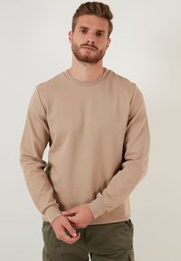 Sudadera beige de mangas largas con cuello redondo y puños acanalados. Tejido suave y texturizado; diseño ajustado; sin logotipos ni patrones visibles.
