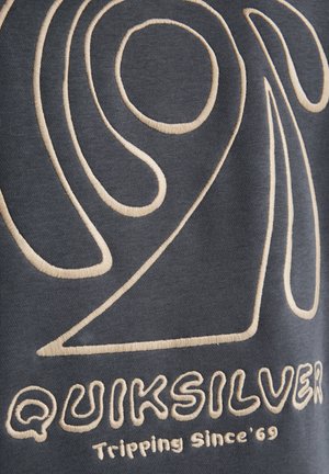 Tummanharmaa kangas, jossa on beigejä kirjailukuvioita. Suunnittelussa korostuvat merkki "Quiksilver" ja iskulause "Tripping Since '69".