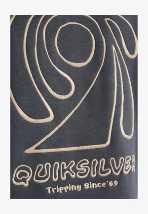 Donkergrijze stof met beige geborduurde patronen. Opvallende merknaam "Quiksilver" en slogan "Tripping Since '69" in het ontwerp verwerkt.