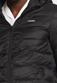 Czarna pikowana kurtka z kapturem, wyposażona w zamknięcie na zamek błyskawiczny, boczne kieszenie oraz logo marki na piersi. Gładka, błyszcząca tekstura.