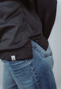 Zbliżenie na osobę noszącą niebieskie jeansy i czarną koszulę z małą białą metką przy krawędzi.