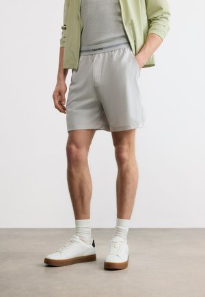 Άνδρας ντυμένος με ανοιχτό γκρι σορτς, γκρι πουκάμισο, πράσινο μπουφάν, λευκές κάλτσες και λευκά αθλητικά παπούτσια με καφέ σόλες, στέκεται σε εσωτερικό χώρο.