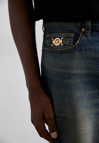Jeans din denim de culoare albastru închis, cu o textură uzată. Prezintă un accent strălucitor de aur pe buzunarul din față și detalii standard de cusătură.