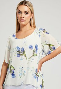 Blusa floreale con vestibilità ampia, caratterizzata da stampe di tulipani blu e piccoli fiori su un tessuto bianco trasparente. Collo rotondo e maniche corte.