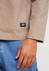 Gros plan d'une personne portant une veste beige Vans avec un jean denim bleu, montrant l'étiquette de la veste près de la poche et de la main droite.