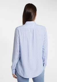 Hellblaues Langarmhemd mit kleinen navyfarbenen Punkten, Knopfleiste und abgerundetem Saum. Der Stoff wirkt strukturiert mit einer weichen Oberfläche.