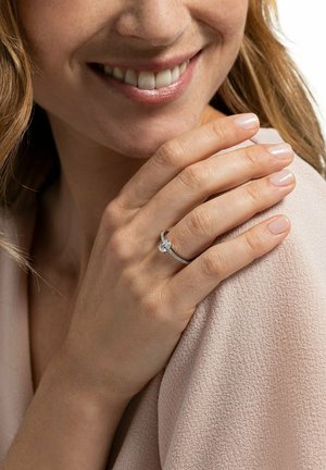 Lächelnde Frau mit hellrosa Nagellack trägt einen silbernen Verlobungsring mit einem auffälligen runden Diamanten am linken Ringfinger.