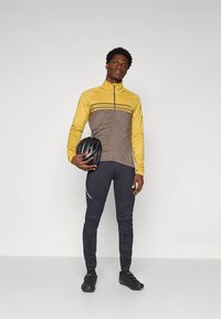 Conjunto de ciclismo com um casaco amarelo e castanho com riscas, combinado com calças pretas. O modelo segura um capacete de ciclismo preto.