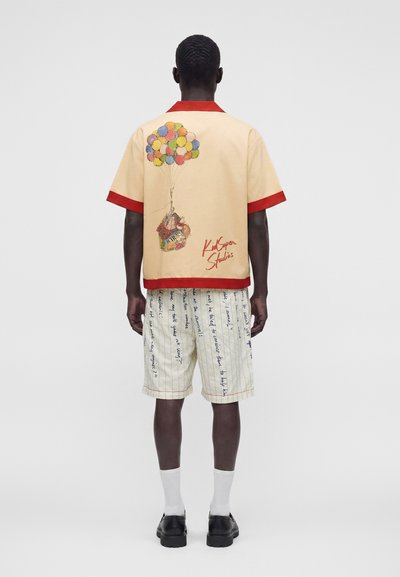 Homme portant une chemise beige à manches courtes avec des bordures rouges et un motif de maison en ballon, associée à un short blanc avec un texte manuscrit bleu.