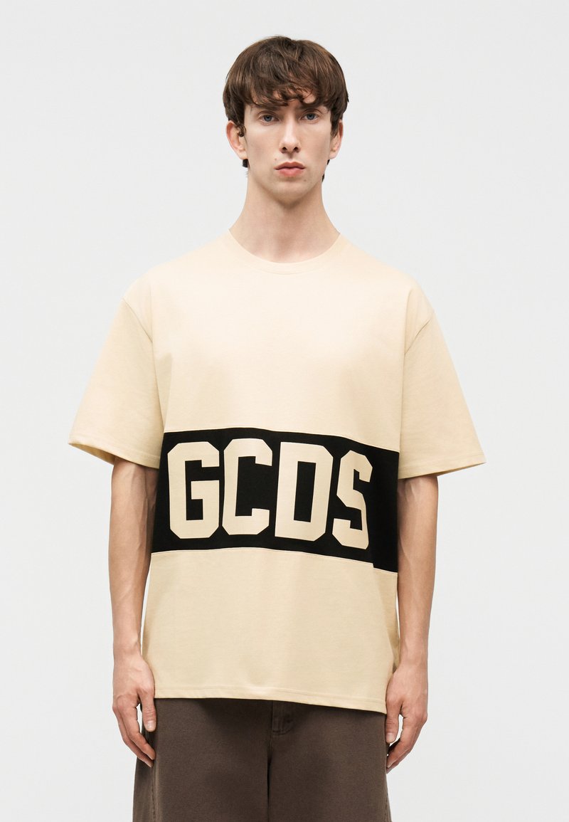 Νεαρός άνδρας που φοράει μπεζ κοντομάνικο πουκάμισο με μια ευρεία μαύρη οριζόντια ρίγα και μεγάλα μπεζ γράμματα "GCDS" στο στήθος.