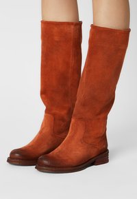 Bottes hautes en daim couleur rouille avec bouts arrondis et talons en bois empilés bas, portées sur des jambes nues.