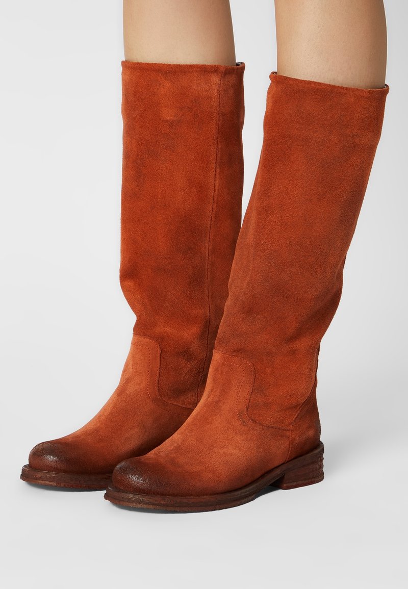 Bottes hautes en daim couleur rouille avec bouts arrondis et talons en bois empilés bas, portées sur des jambes nues.